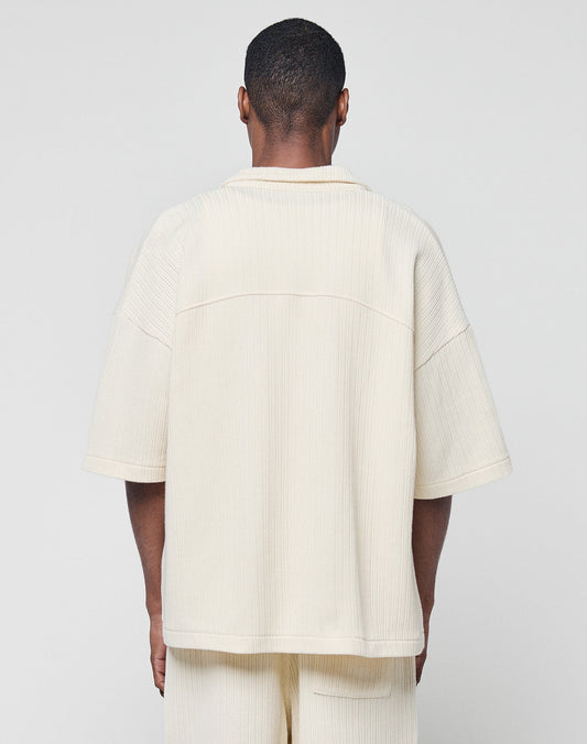 A person faces away from the camera, wearing the LIVE FAST DIE YOUNG Basic Knit Shirt in a cream, oversize fit with short sleeves and Hemdkragen, paired with matching pants against a plain light background.