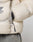 Close-up of a hand partially tucked into the zippered side pocket of a light beige Live Fast Puffer Jacket by LIVE FAST DIE YOUNG, featuring a visible logo on the left and paired with grey pants.
