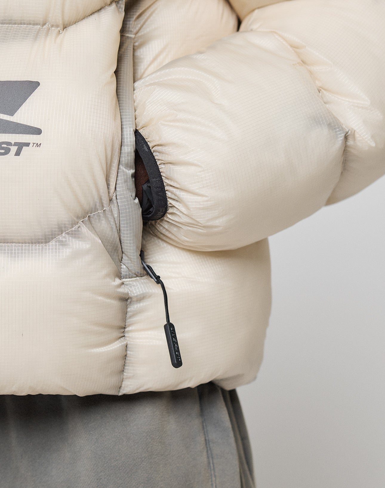 Close-up of a hand partially tucked into the zippered side pocket of a light beige Live Fast Puffer Jacket by LIVE FAST DIE YOUNG, featuring a visible logo on the left and paired with grey pants.