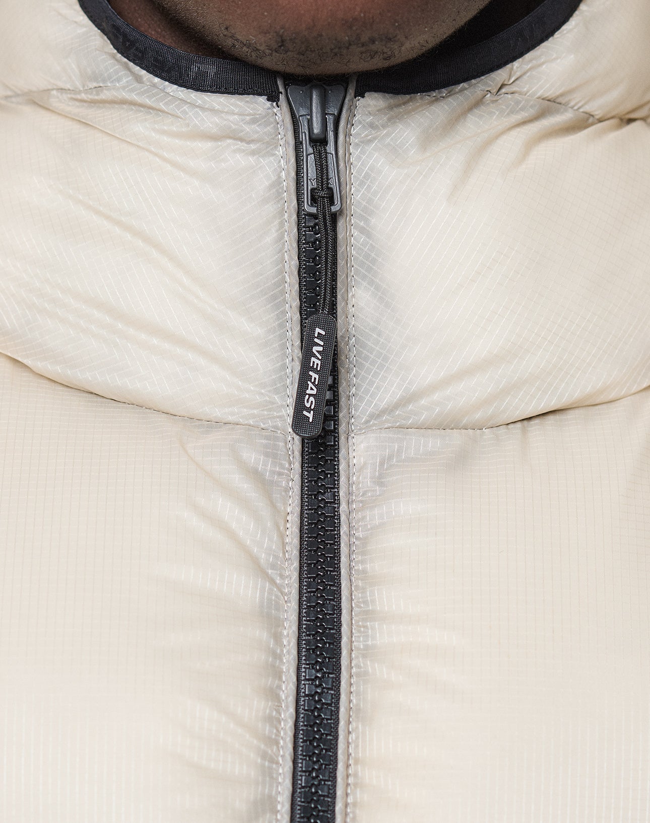 Close-up of a person wearing the LIVE FAST DIE YOUNG Live Fast Puffer Jacket, featuring a black zipper with a "LIVE FAST" pull tab, focused on the upper chest and neck area.