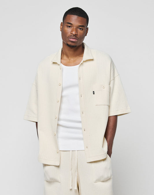 A man wears a LIVE FAST DIE YOUNG Basic Knit Shirt in cream over a white tank top with matching shorts, standing against a light gray background and looking at the camera with a neutral expression.