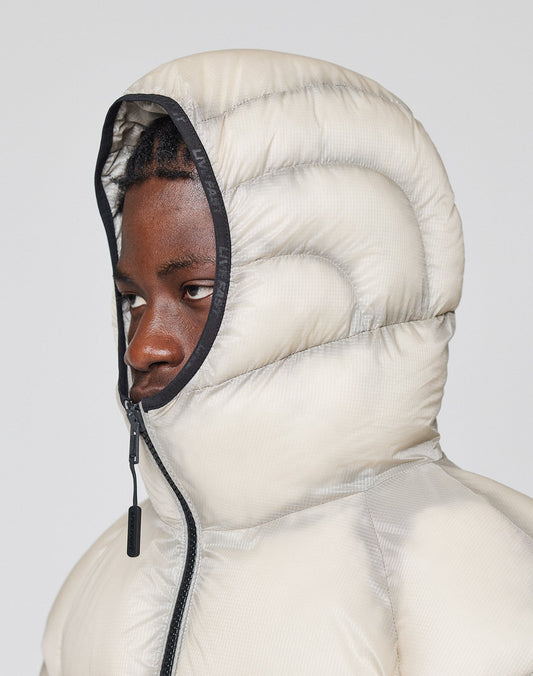A person wears the Live Fast Puffer Jacket by LIVE FAST DIE YOUNG in oversized light beige nylon, with the hood fully zipped up to cover most of their head except the face. The background is plain and neutral.