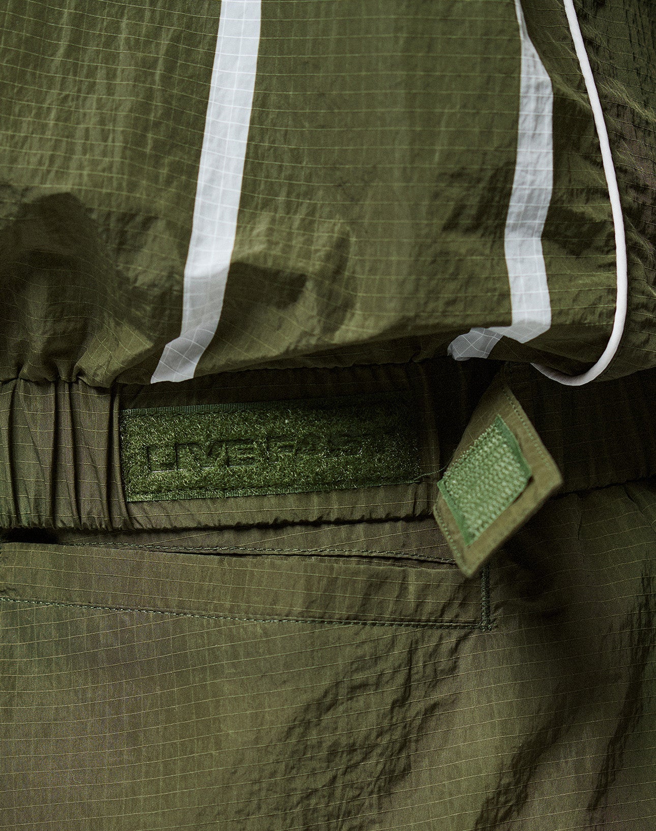Close-up of an olive green LFDY Nylon Trackjacket by LIVE FAST DIE YOUNG, featuring crinkled fabric, white reflective stripes, Velcro strap, elastic hood drawstring, embossed patch detail, and an oversize fit for comfort.