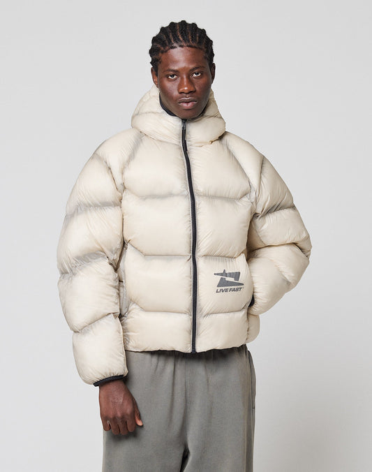 A person stands against a plain light background wearing the LIVE FAST DIE YOUNG Live Fast Puffer Jacket in beige with a hood, black zipper, front pocket patch reading "LIVE FAST," gray pants, and a reflective print.