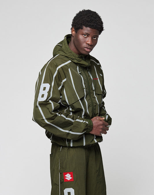 A young man with short curly hair wears the olive green LFDY Nylon Trackjacket by LIVE FAST DIE YOUNG, featuring stripes, front and back print, and the number 8 on the sleeve. He poses confidently against a plain light background.
