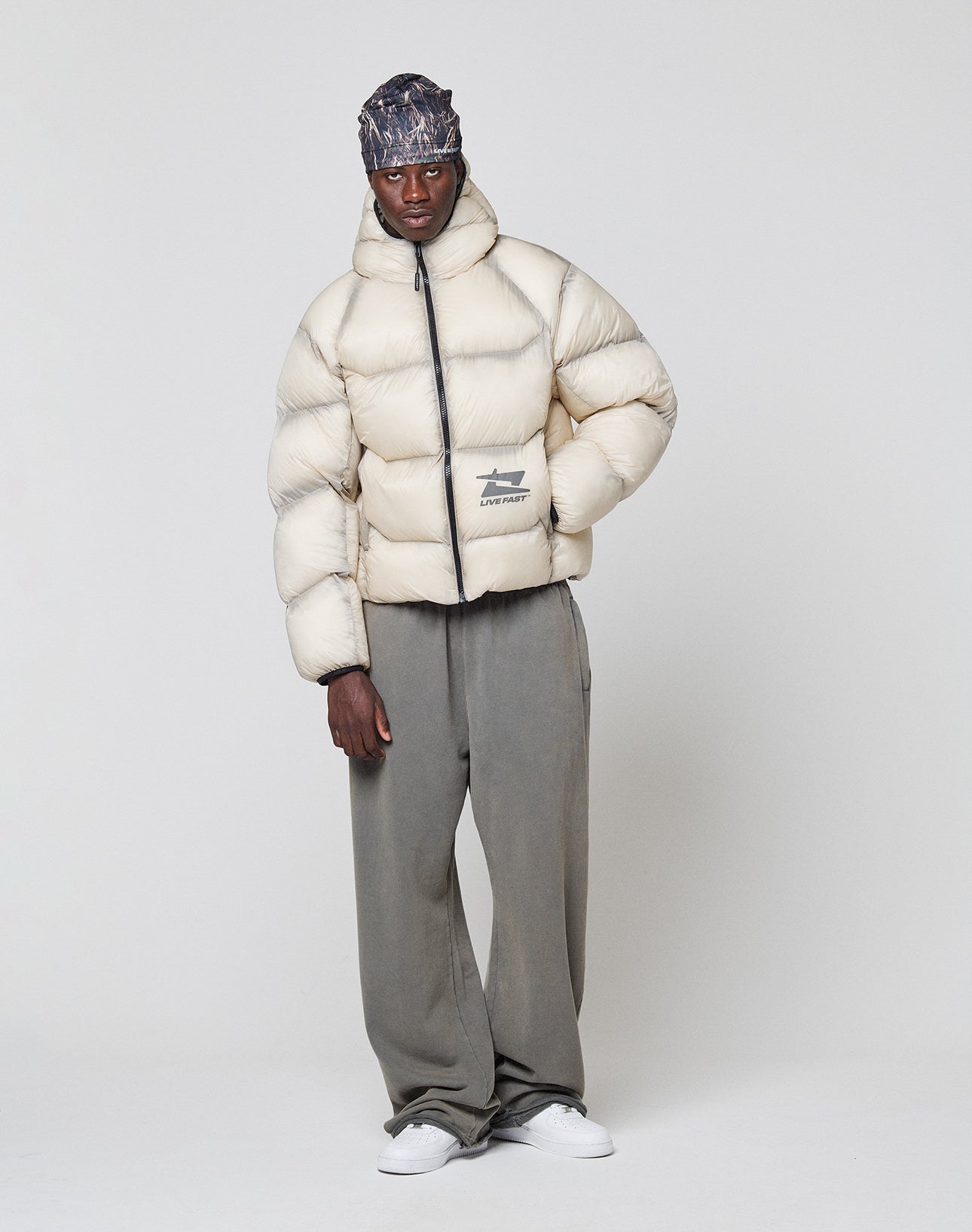 A man stands against a plain background wearing the LIVE FAST DIE YOUNG Live Fast Puffer Jacket in cream, paired with loose gray pants, white sneakers, and a patterned headscarf. He faces forward with a relaxed posture and neutral expression.