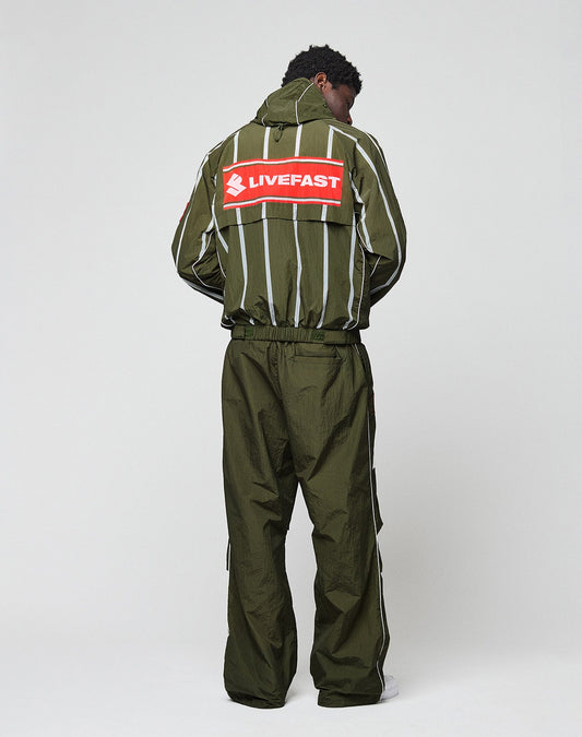 A person wears the LFDY Nylon Trackjacket by LIVE FAST DIE YOUNG—an olive green, oversize-fit jacket with white stripes, a stylized running figure, and a bold red LIVEFAST backprint for standout sporty style.