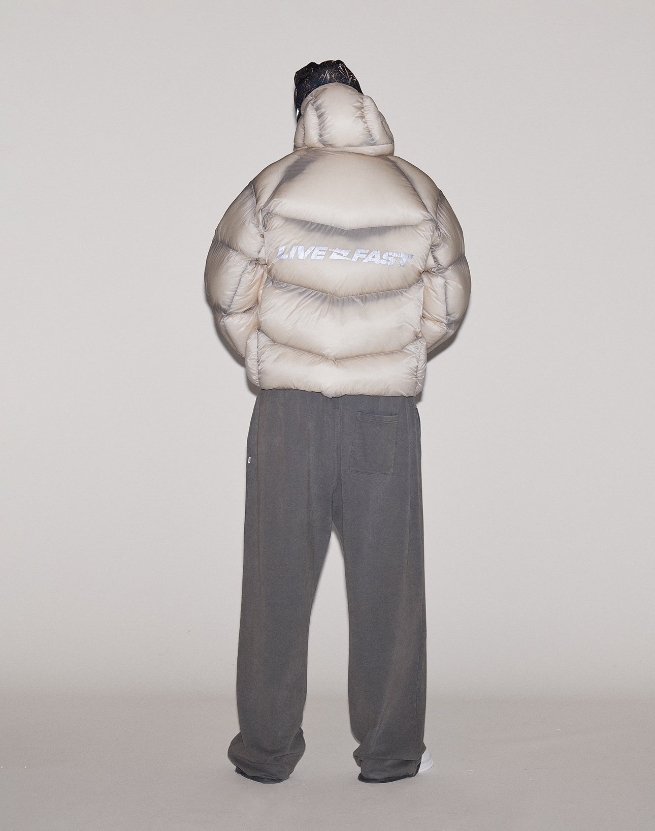 A person stands against a plain light background, wearing the LIVE FAST DIE YOUNG Live Fast Puffer Jacket in light beige with an oversized fit and reflective "LIVE FAST" print on the back, paired with loose dark pants and a head covering.