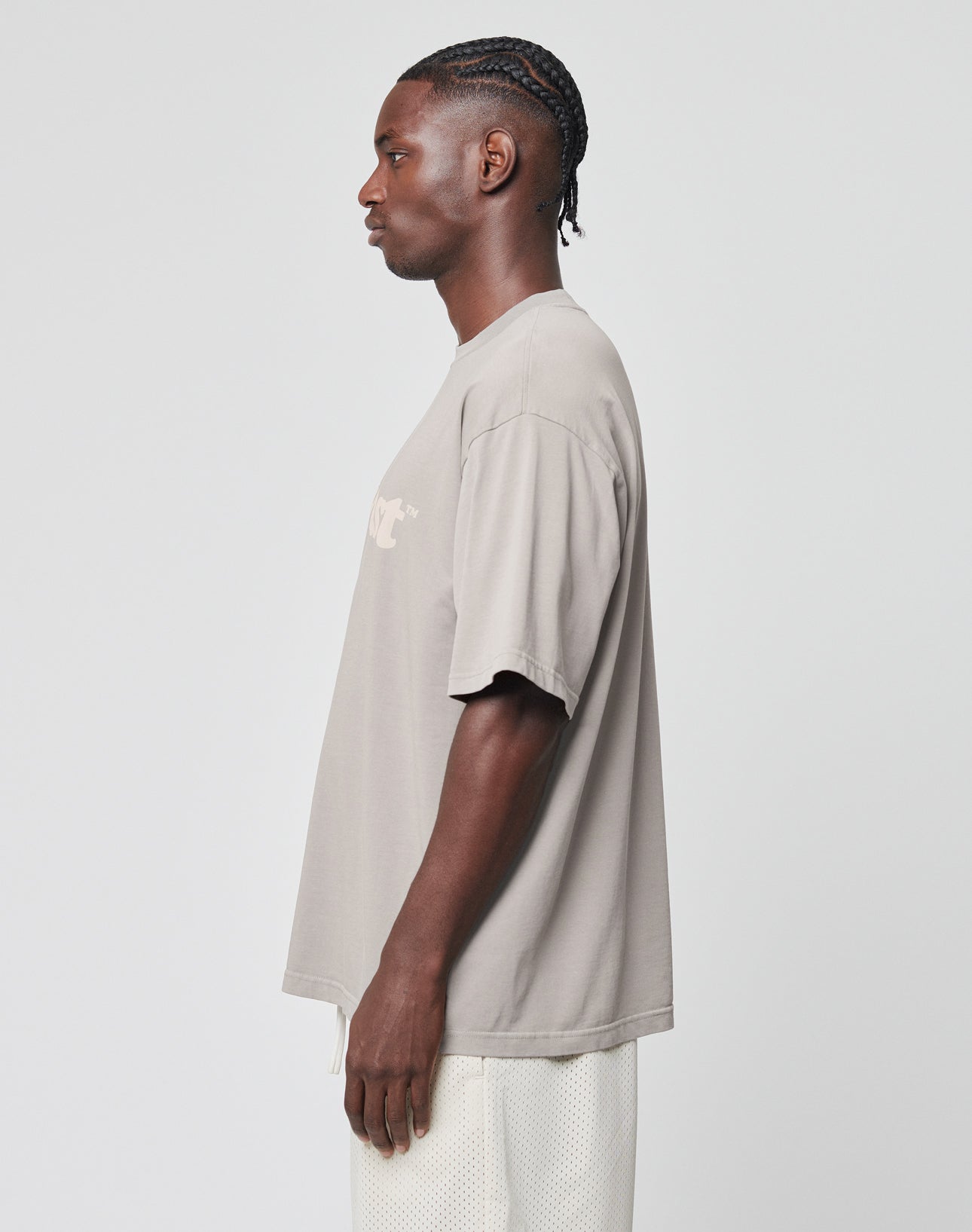 A man with braided hair stands in profile against a plain background, wearing a LIVE FAST DIE YOUNG Basic Tee in oversize gray 100% Baumwolle and white mesh shorts.