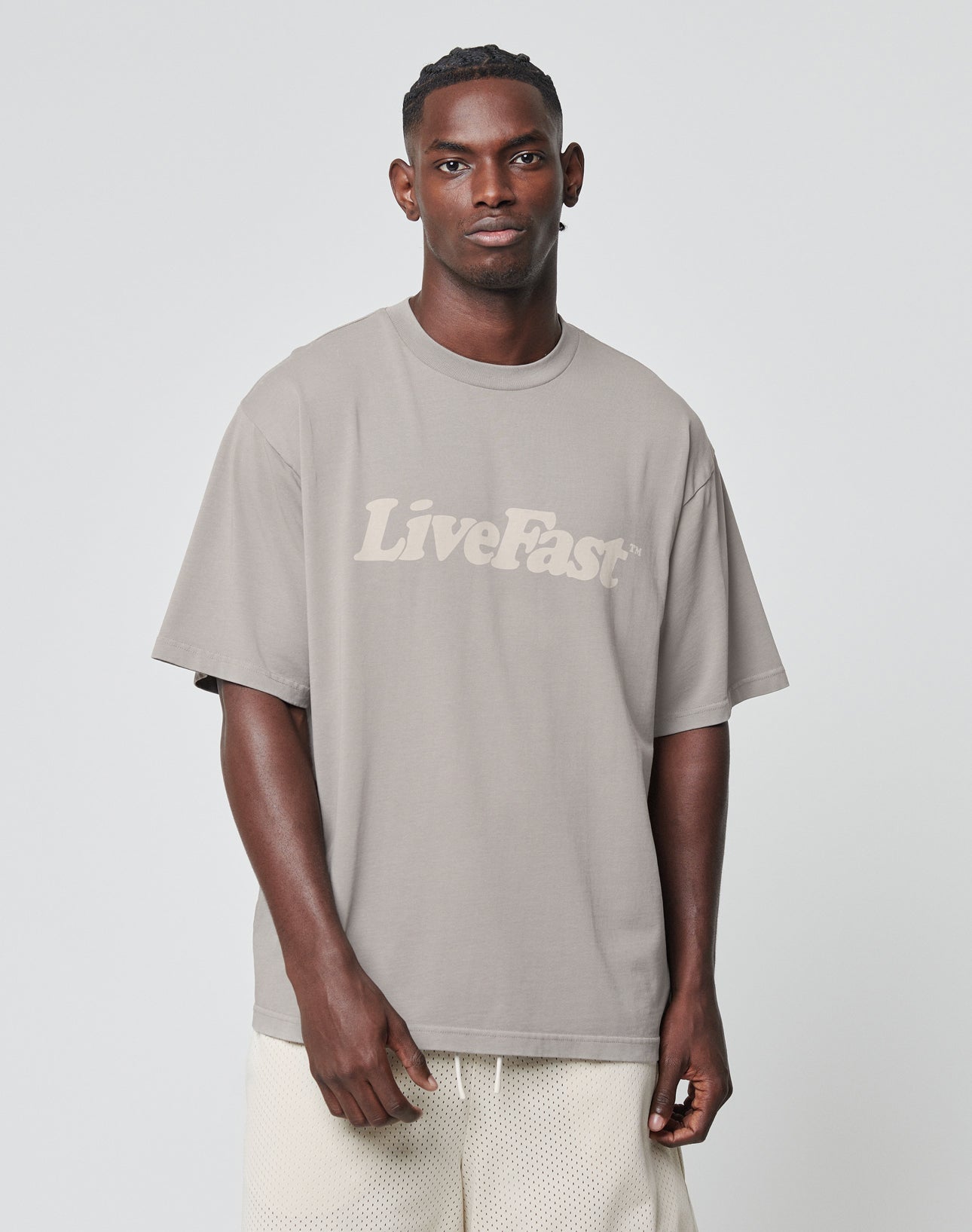 A man stands against a plain background wearing a taupe Oversize Fit Basic Tee from LIVE FAST DIE YOUNG with a vintage wash and bold LiveFast print, paired with light-colored mesh shorts.
