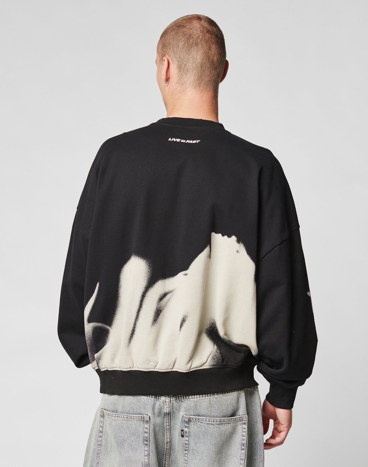A person with short hair, seen from behind, wears the LFDY Mystic Sweater by LIVE FAST DIE YOUNG—black with white abstract patterns and LIVE FAST text on the upper back—paired with light gray Oversize Fit jeans against a plain background.