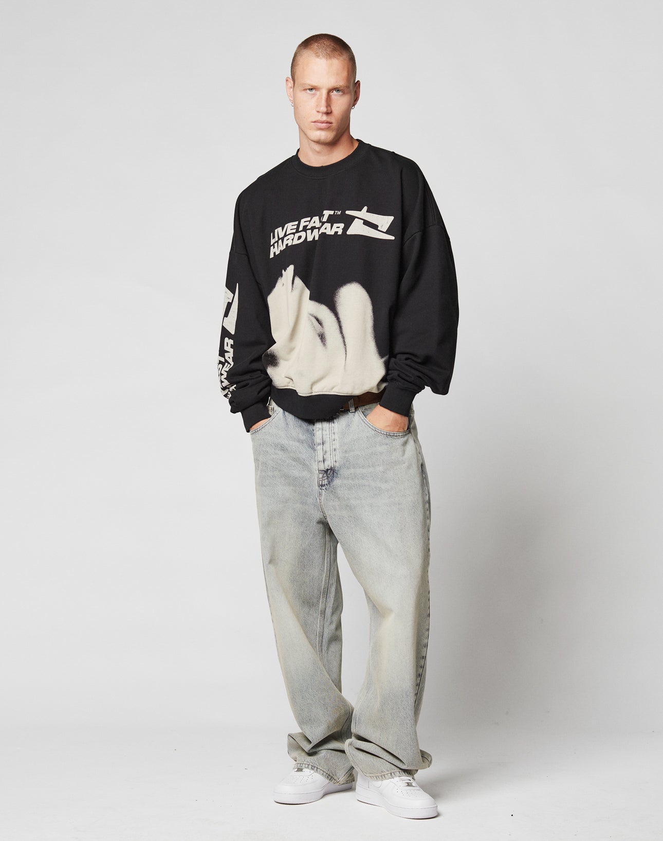 A man with short hair wears the LIVE FAST DIE YOUNG LFDY Mystic Sweater, an oversized black graphic sweatshirt, paired with baggy light-wash jeans and white sneakers. He stands hands-in-pockets against a plain light gray background.
