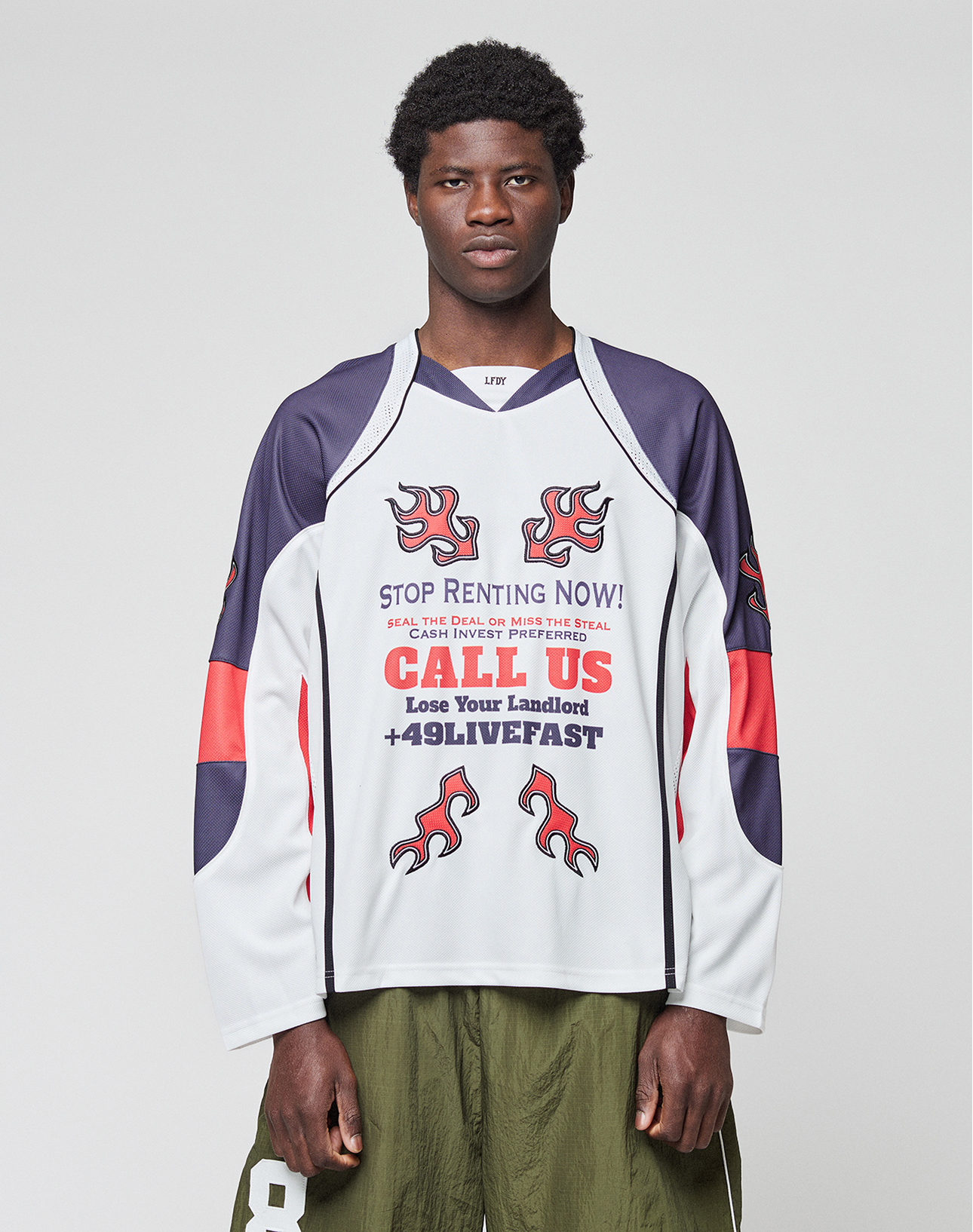 A young man stands facing forward wearing the LIVE FAST DIE YOUNG LFDY Call Us Jersey, an oversize white long-sleeve shirt with blue and red accents, flame graphics, and bold text. He pairs it with olive green shorts. | model