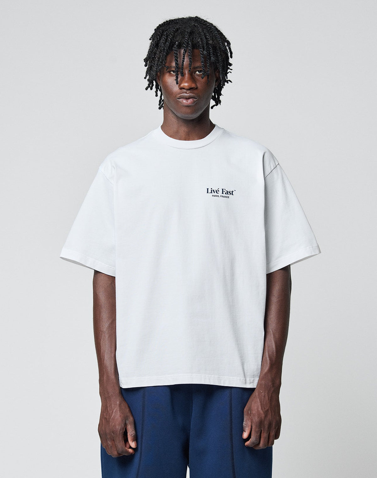 A person with medium-length dreadlocks wears the LFDY Paris Tee by LIVE FAST DIE YOUNG, an oversized white cotton shirt with front and back print, paired with dark blue pants, standing against a plain light gray background.