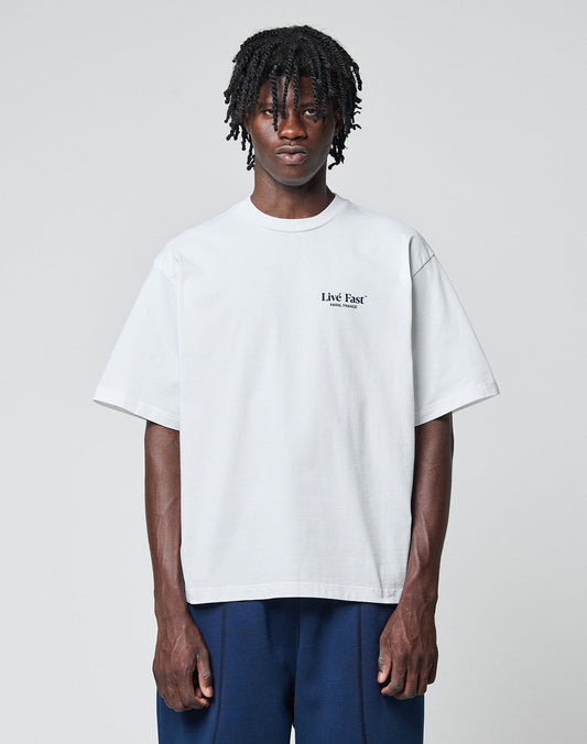 A person with medium-length dreadlocks wears the LFDY Paris Tee by LIVE FAST DIE YOUNG, an oversized white cotton shirt with front and back print, paired with dark blue pants, standing against a plain light gray background.