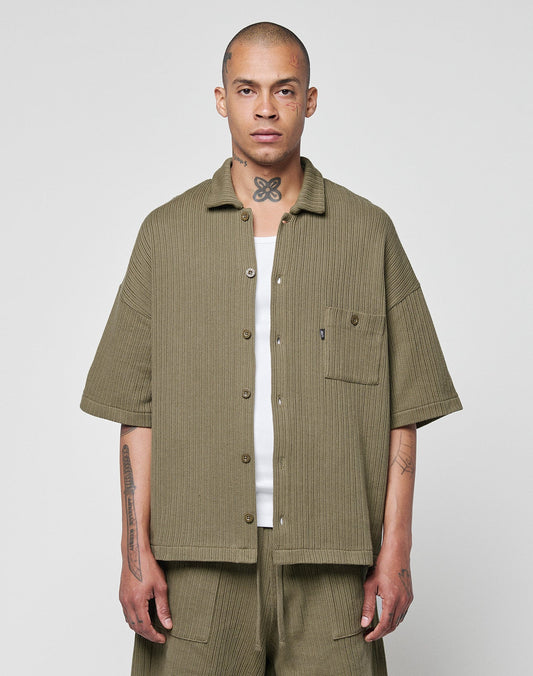 A person with a shaved head and visible tattoos stands facing forward, wearing the LIVE FAST DIE YOUNG Basic Knit Shirt in olive green with a Hemdkragen and Brusttasche, paired with wide-legged matching shorts against a plain light gray background.