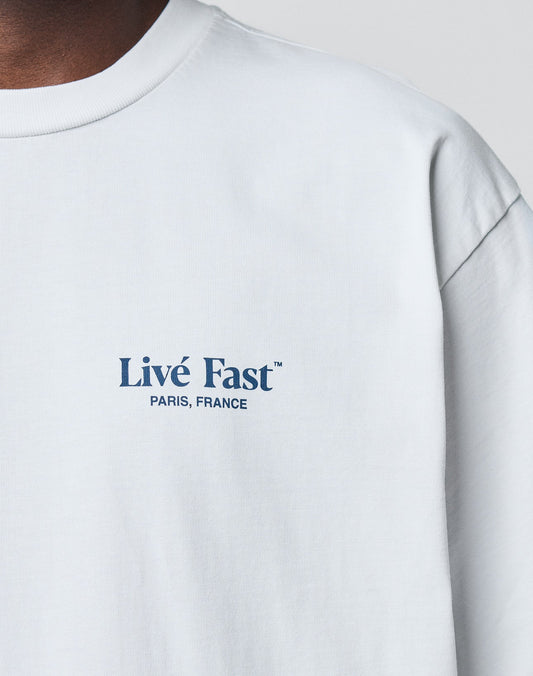 A close-up of the LIVE FAST DIE YOUNG LFDY Paris Tee in white Baumwolle shows Livé Fast™ PARIS, FRANCE in blue on the left chest. Only part of the neck and shoulder are visible, emphasizing its Oversize Fit.