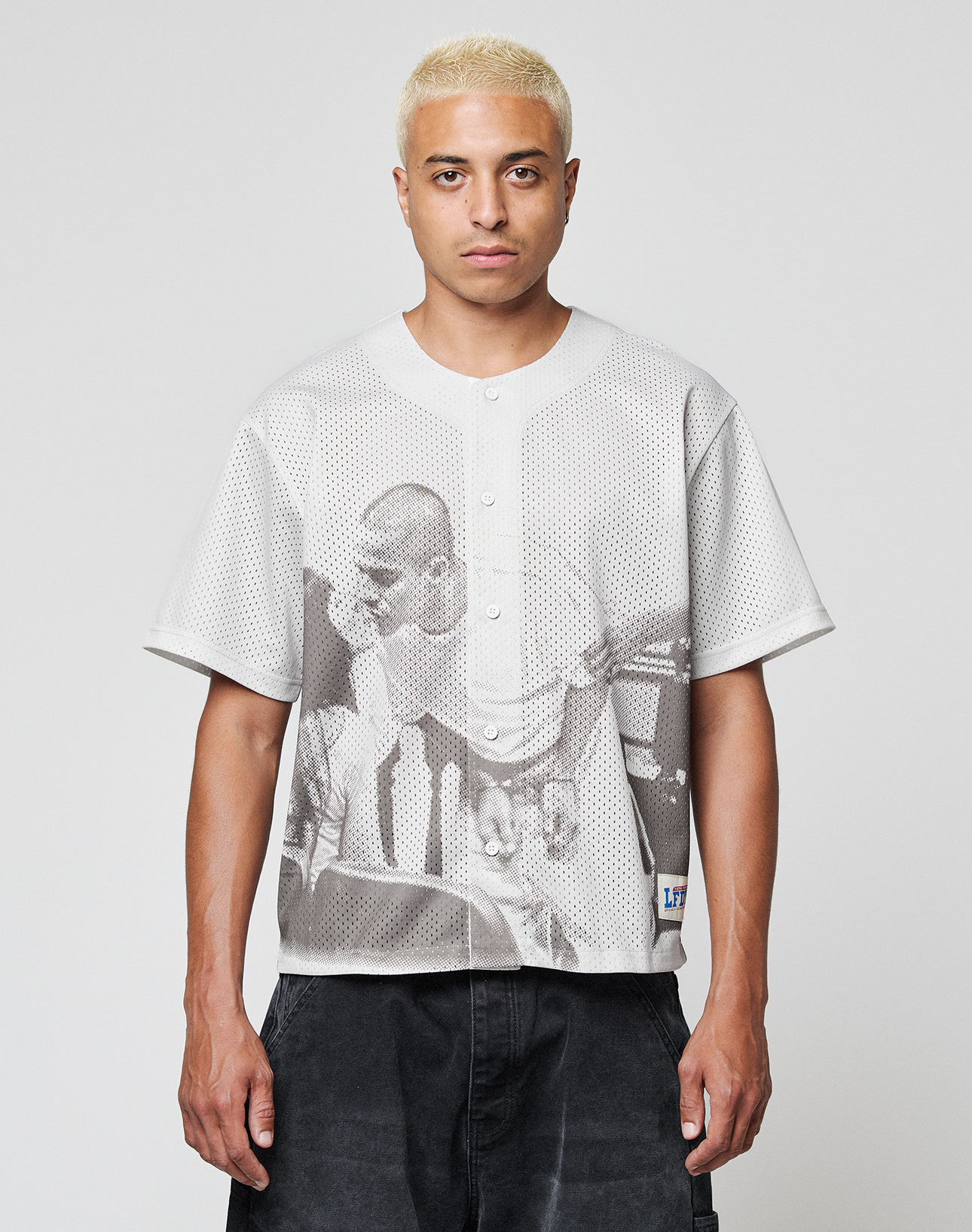 A person with short bleached blond hair wears the LIVE FAST DIE YOUNG LFDY Courtroom Jersey, an oversized light gray mesh button-up featuring a large black-and-white print of two men on the front, standing against a plain light background. | model