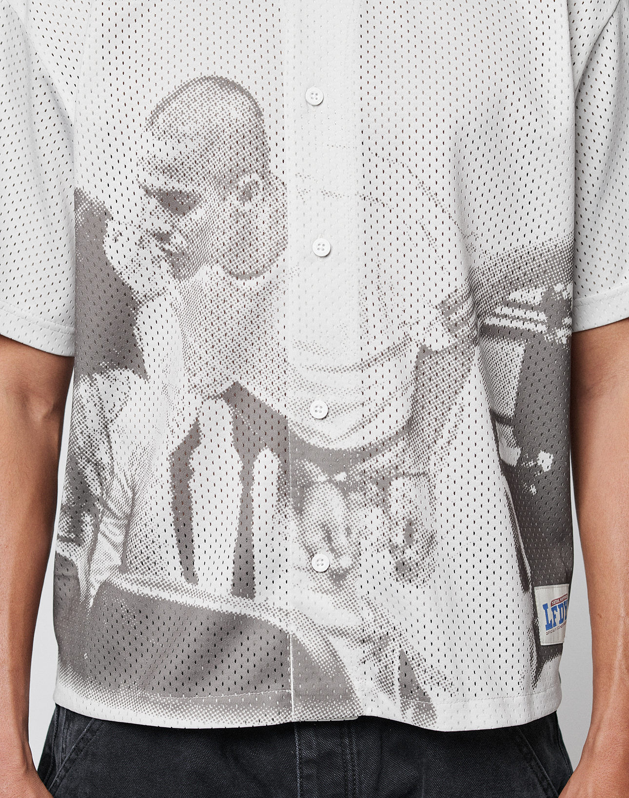 A close-up of a person wearing the LFDY Courtroom Jersey by LIVE FAST DIE YOUNG, an oversize fit white mesh shirt with a grayscale all-over print of two people, paired with black pants.