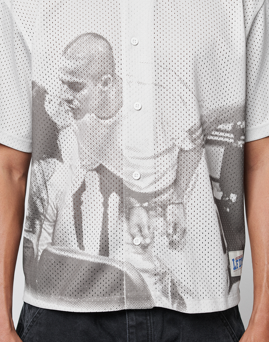 A close-up of a person wearing the LFDY Courtroom Jersey by LIVE FAST DIE YOUNG, an oversize fit white mesh shirt with a grayscale all-over print of two people, paired with black pants.