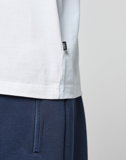 Close-up of a person wearing the LFDY Paris Tee by LIVE FAST DIE YOUNG in white, featuring a small black tag on the side seam, styled with blue pants with a visible zipper pocket. The background is plain and light-colored.