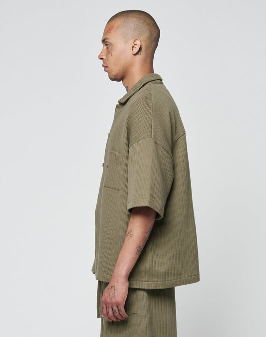 A person with a shaved head stands in profile against a plain background, wearing LIVE FAST DIE YOUNGs olive Basic Knit Shirt and matching pants.