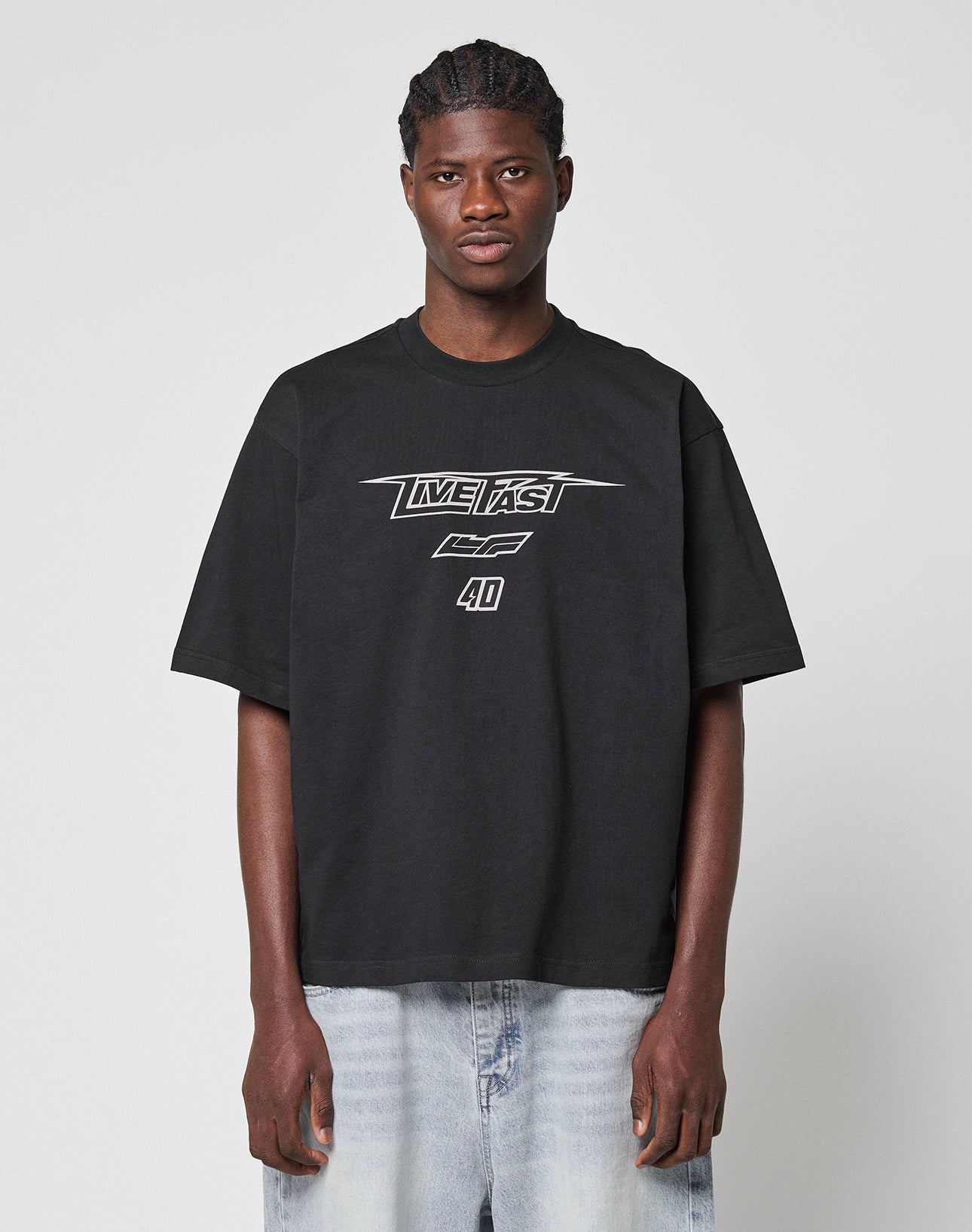 A person with braided hair wears the LFDY Triple Racing Tee by LIVE FAST DIE YOUNG—featuring a multilogo front print and made from 100% Baumwolle—paired with light blue jeans, standing against a plain light gray background.