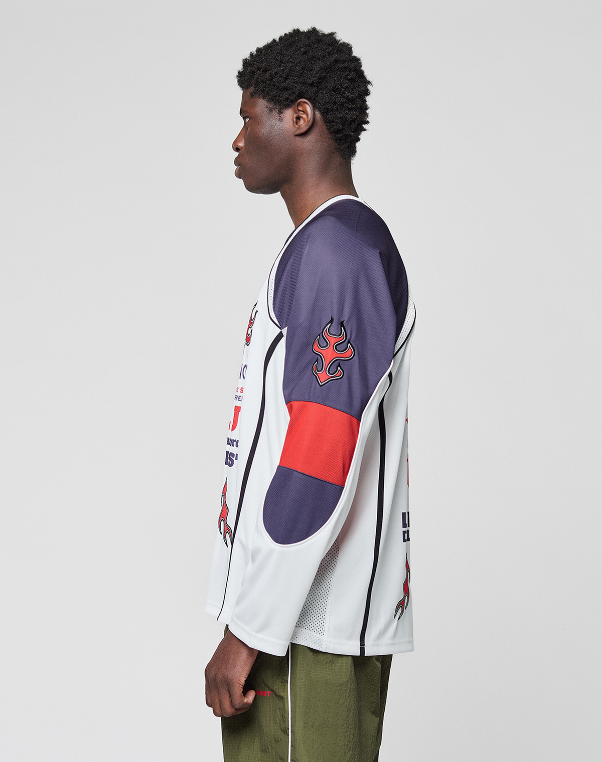 A person stands in profile facing left, wearing the LIVE FAST DIE YOUNG LFDY Call Us Jersey—an oversize white polyester long-sleeve shirt with navy and red flame graphics on the arm—and olive green pants against a plain light gray background.