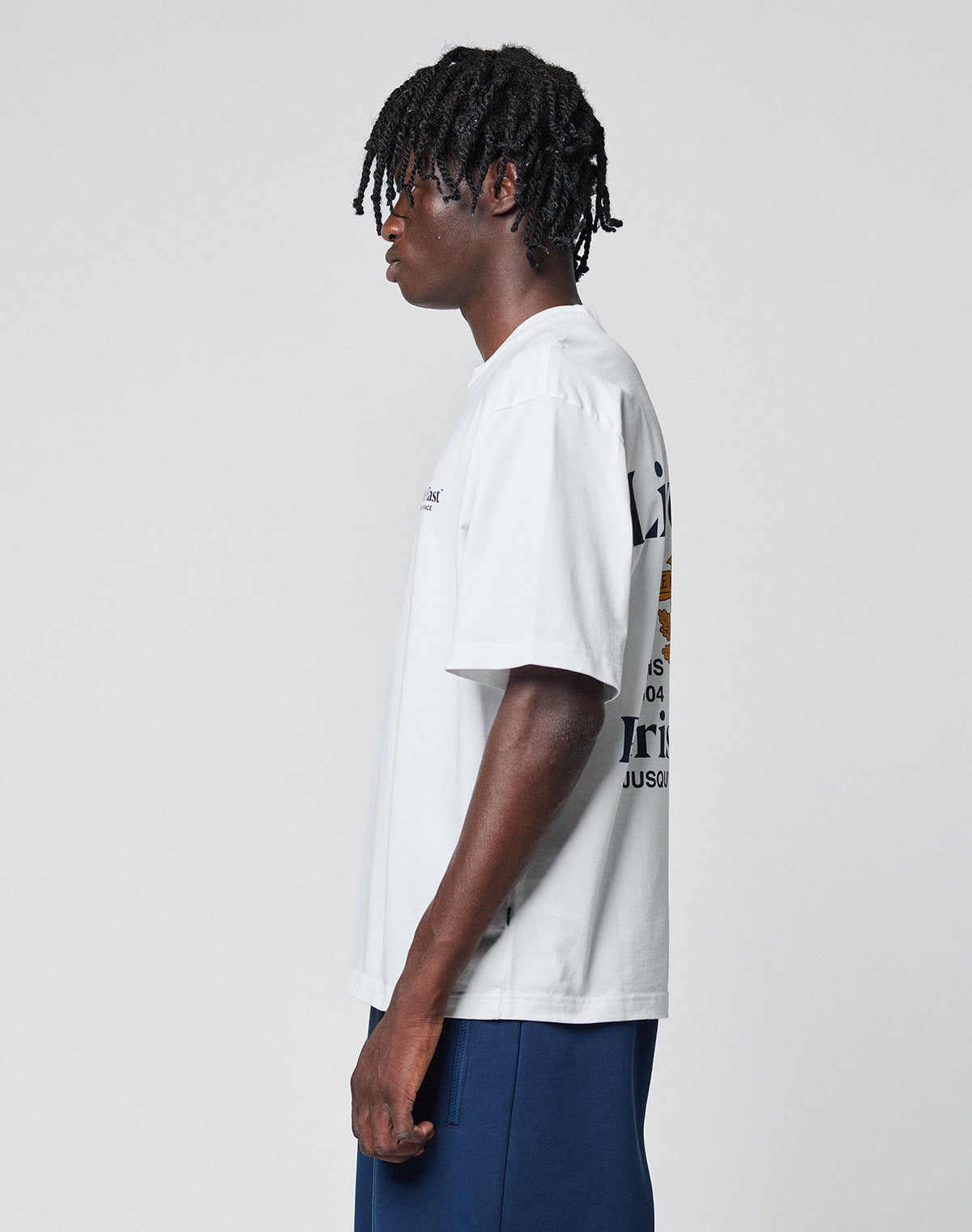 A man with short dreadlocks stands in profile wearing a white LFDY Paris Tee by LIVE FAST DIE YOUNG, featuring front and back print, paired with navy blue pants against a plain light gray background.