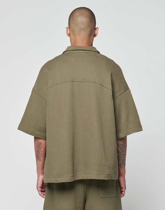 A person with a shaved head is seen from behind wearing an olive green LIVE FAST DIE YOUNG Basic Knit Shirt, an oversize ribbed short-sleeve top with Hemdkragen and Brusttasche. Their tattooed arms stand out against a plain light background.