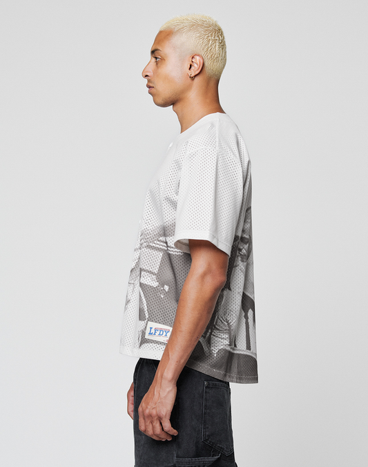 A person with short, bleached blond hair stands in profile against a plain background, wearing an oversize fit LIVE FAST DIE YOUNG LFDY Courtroom Jersey and dark shorts.