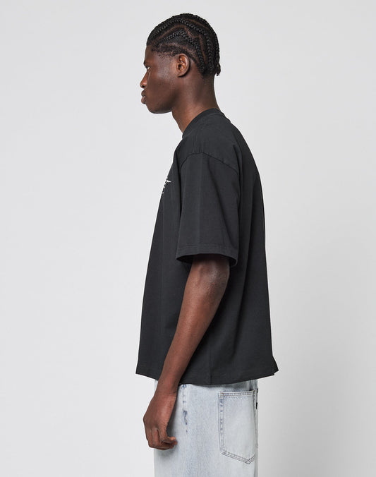 A person with braided hair stands in profile, wearing a loose black LIVE FAST DIE YOUNG LFDY Triple Racing Tee and light blue jeans against a plain light gray background.