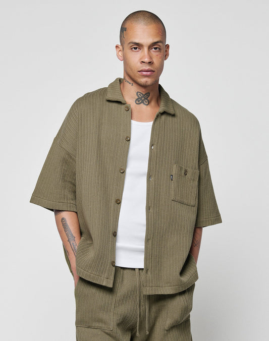 A person with a shaved head and tattoos wears the LIVE FAST DIE YOUNG Basic Knit Shirt in olive green, styled oversized over a white tank and matching pants, standing against a plain light gray background.