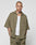 A person with a shaved head and tattoos wears the LIVE FAST DIE YOUNG Basic Knit Shirt in olive green, styled oversized over a white tank and matching pants, standing against a plain light gray background.