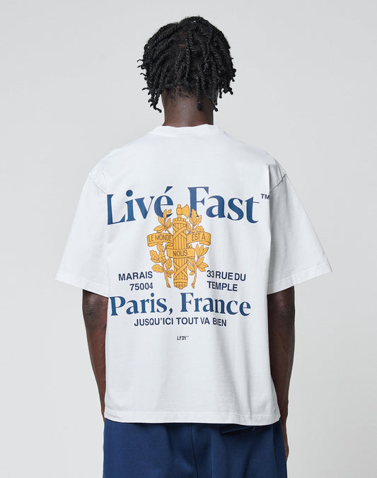 A person wearing the LFDY Paris Tee by LIVE FAST DIE YOUNG, an oversized white cotton T-shirt with blue and yellow front and back graphics reading “Live Fast™ Paris, France” and French text, stands against a plain light background. | model