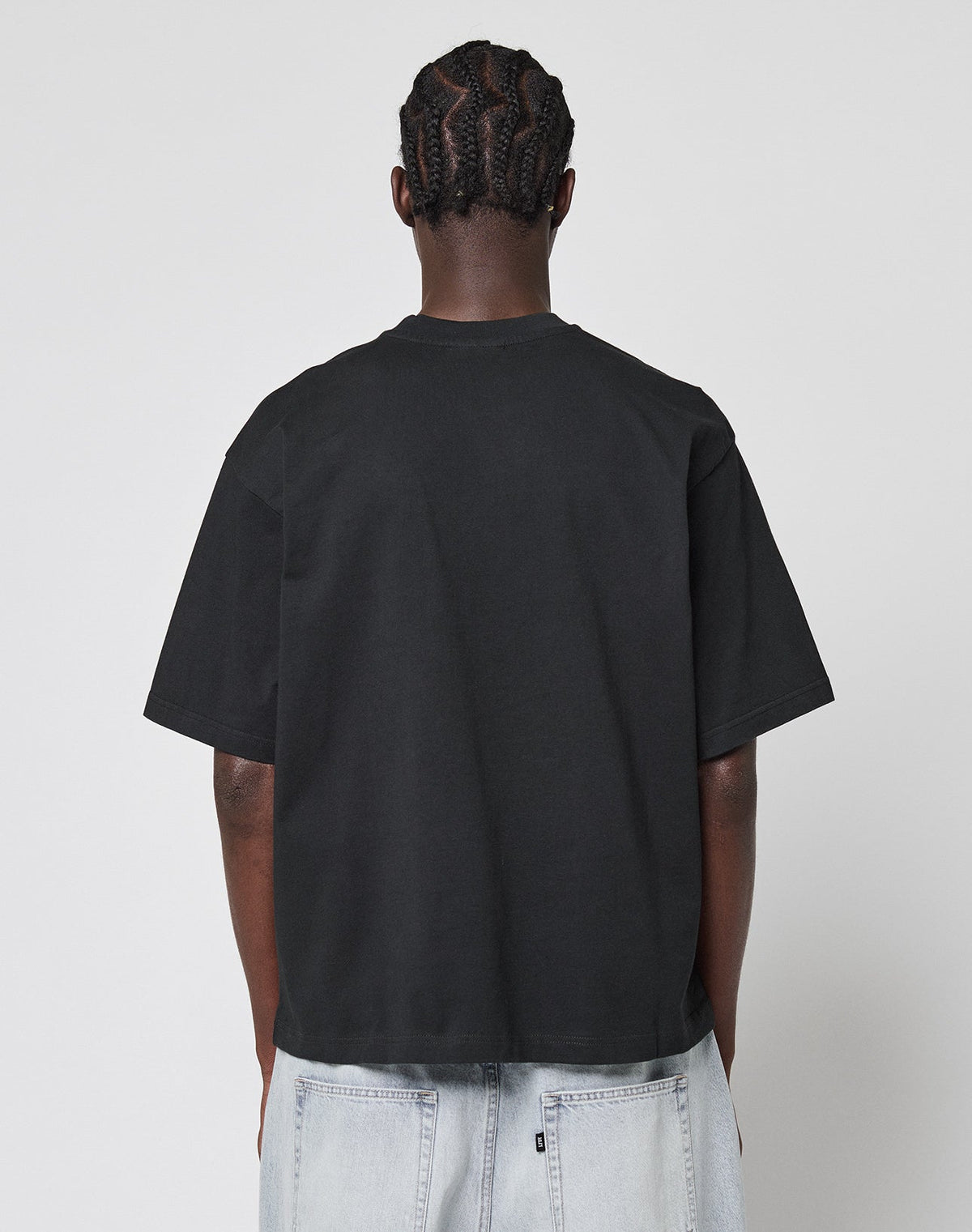 A person with braided hair stands with their back to the camera, wearing a LIVE FAST DIE YOUNG LFDY Triple Racing Tee in black and light-wash denim jeans against a plain white background.