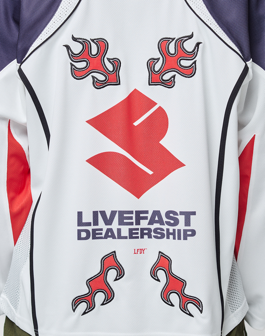 The LFDY Call Us Jersey by LIVE FAST DIE YOUNG is a white polyester oversize-fit sports jersey with a bold red abstract logo, four flame graphics, and "LIVEFAST DEALERSHIP" in purple. Red and dark blue accents highlight the sides and shoulders.