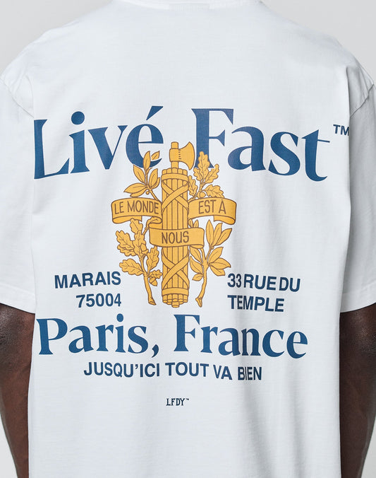Someone wears the LFDY Paris Tee by LIVE FAST DIE YOUNG—an oversize white cotton shirt with blue Livé Fast and Paris, France text, ornate yellow cross graphics, French words, and 33 Rue du Temple, Marais 75004 on front and back.
