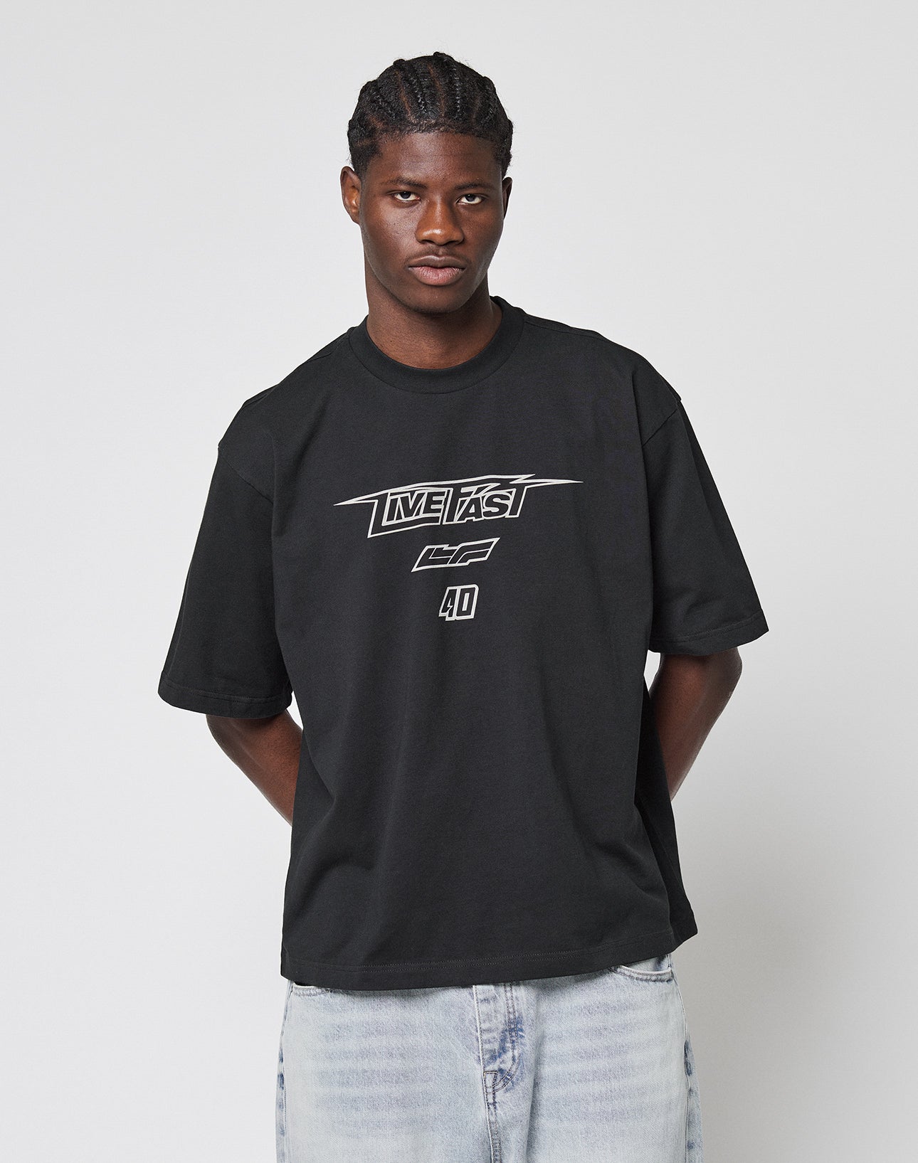 A man in a black LFDY Triple Racing Tee by LIVE FAST DIE YOUNG, featuring multilogo frontprint and “LIVE FAST” text, made from 100% Baumwolle, with light blue jeans stands against a plain white background, hands behind his back.