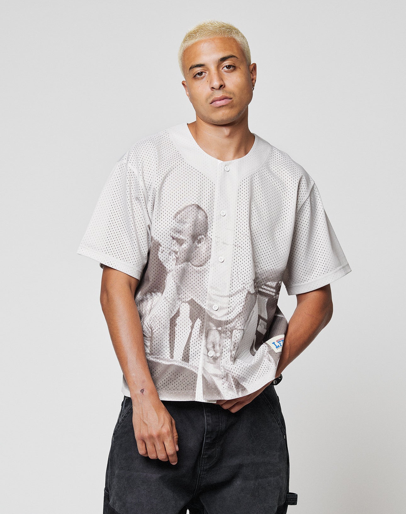 A person with short blond hair wears the LIVE FAST DIE YOUNG LFDY Courtroom Jersey—an oversized, light mesh shirt with a large basketball player print—paired with black pants, against a plain gray background.