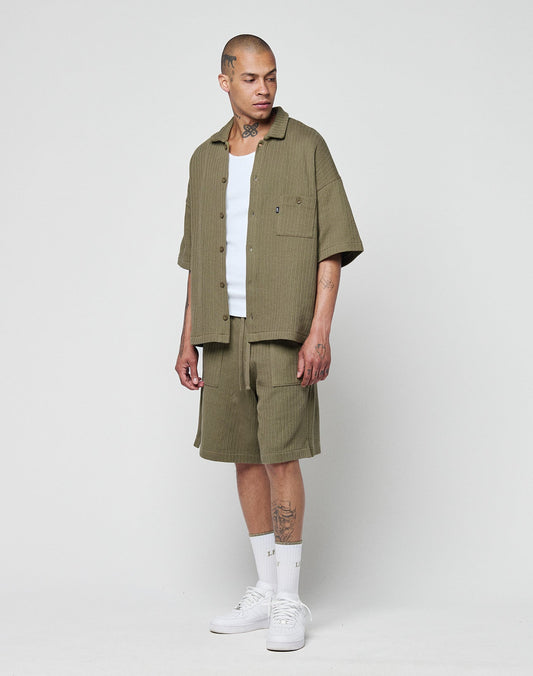 A person with tattoos and a neutral expression wears the LIVE FAST DIE YOUNG Basic Knit Shirt in olive green (short-sleeve, oversize fit with Hemdkragen and Brusttasche), matching shorts, a white tank top, white socks, and white sneakers.