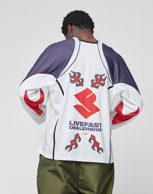 A person faces away wearing the LIVE FAST DIE YOUNG LFDY Call Us Jersey, a white, navy, and red long-sleeve with flame graphics and "LIVEFAST DEALERSHIP" on the back, in an Oversize Fit, paired with olive green pants.