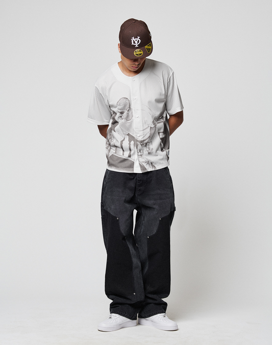 A man stands against a plain white background, looking down with his hands behind his back, wearing the LFDY Courtroom Jersey by LIVE FAST DIE YOUNG, loose black jeans, an oversize brown cap with yellow graphics, and white sneakers.