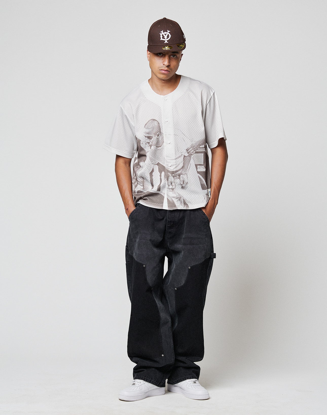 A young man stands against a plain background, wearing a brown cap, sunglasses, a light grey graphic shirt, white sneakers, and LIVE FAST DIE YOUNG LFDY Carpenter Pants—baggy fit black jeans made from 100% Baumwolle—with hands in his pockets.