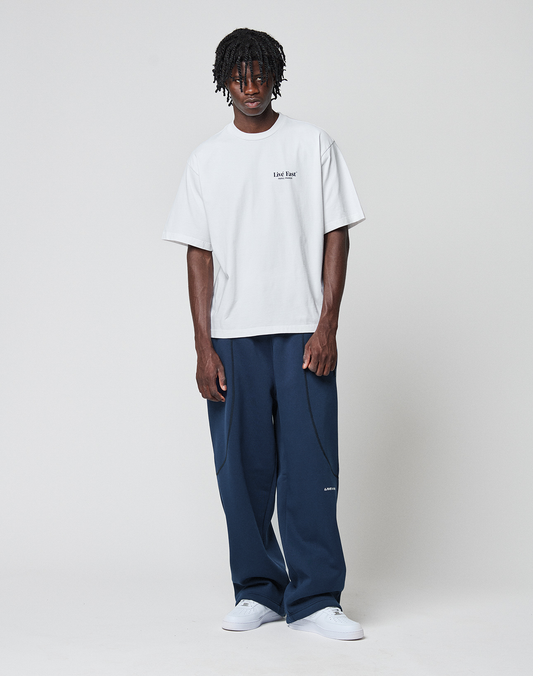 A man with short dreadlocks stands against a plain background, wearing a loose white logo T-shirt, LIVE FAST DIE YOUNG LFDY Active Fleece Jogger pants in navy blue, and white sneakers. He faces forward with arms relaxed by his sides.