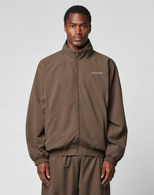 A person faces the camera against a plain gray background, wearing the brown polyester LFDY Trackjacket by LIVE FAST DIE YOUNG with an oversize fit and LIVE FAST text on the left chest, paired with matching pants.