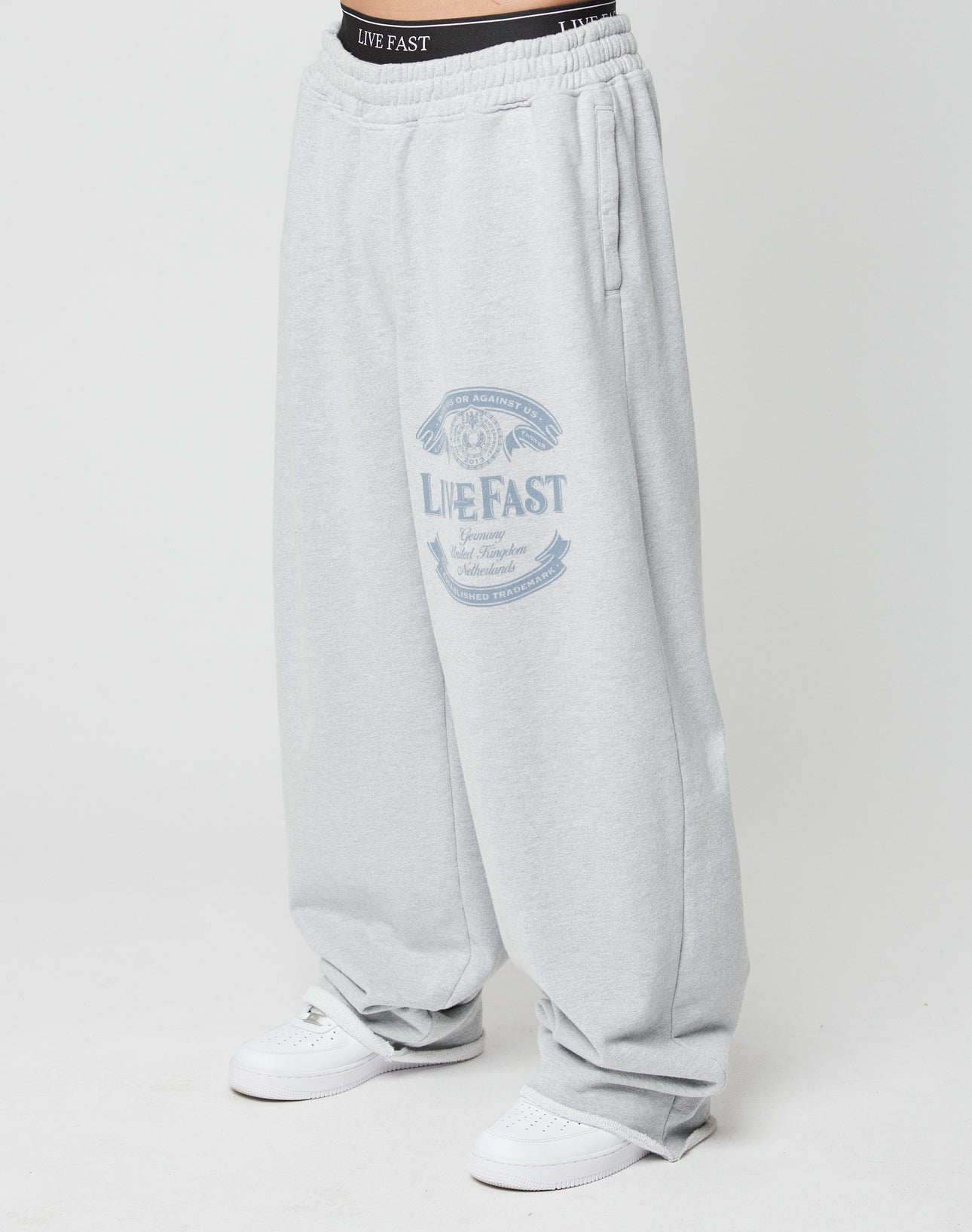 A person wears Loyal Wide Jogger sweatpants by LIVE FAST DIE YOUNG in light gray, featuring a wide leg, elastic waistband, and graphic on the upper thigh. Black underwear is visible above the pants, paired with white sneakers.