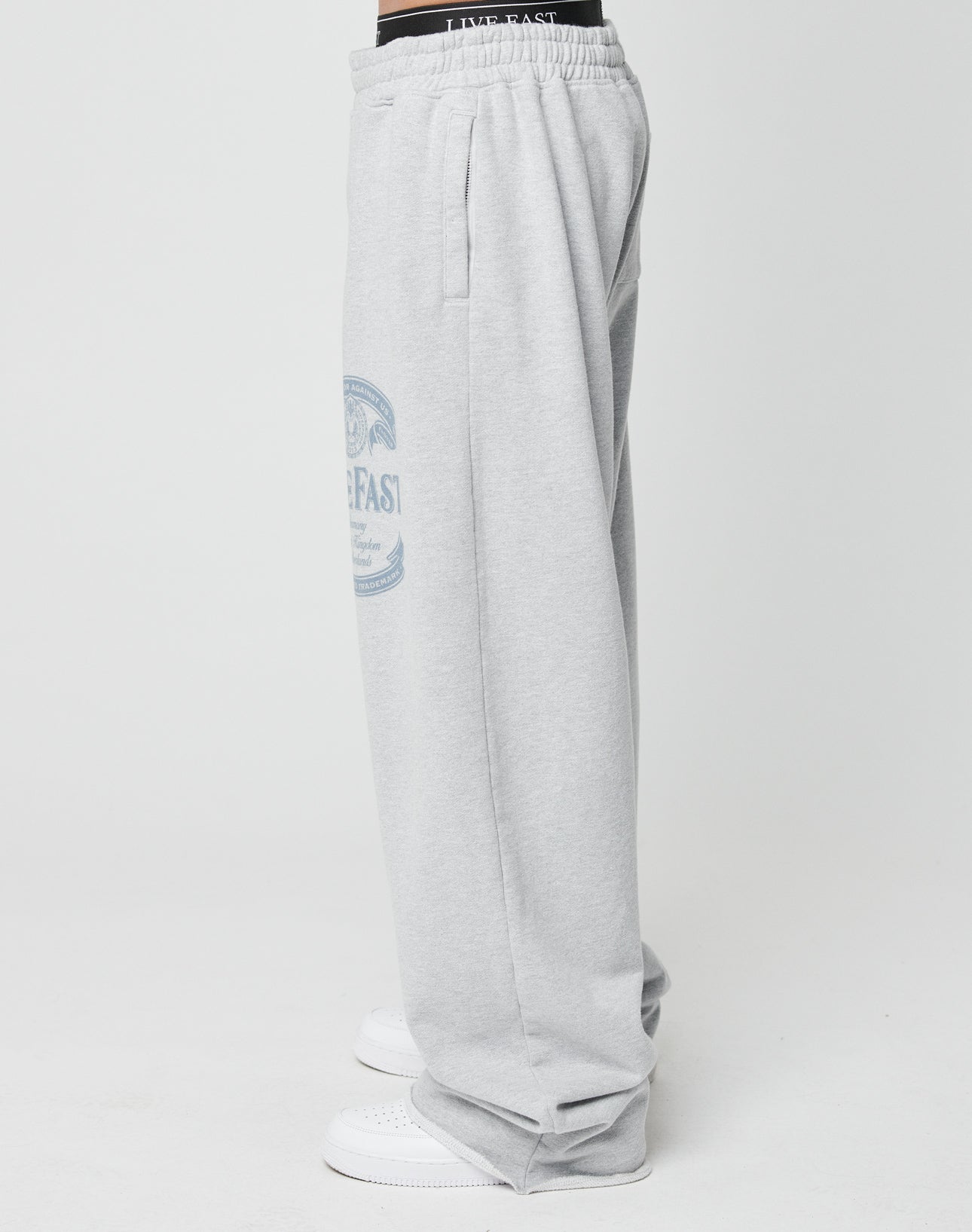 A person models LIVE FAST DIE YOUNG’s Loyal Wide Jogger—light gray with wide leg and elastic waistband, blue graphic on the thigh—paired with white sneakers and a visible black waistband, standing sideways against a white background.