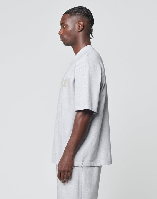 A man with braided hair stands in profile, wearing an oversized light gray LIVE FAST DIE YOUNG Basic Tee and matching shorts made from a soft cotton-polyester blend, set against a plain, light background.
