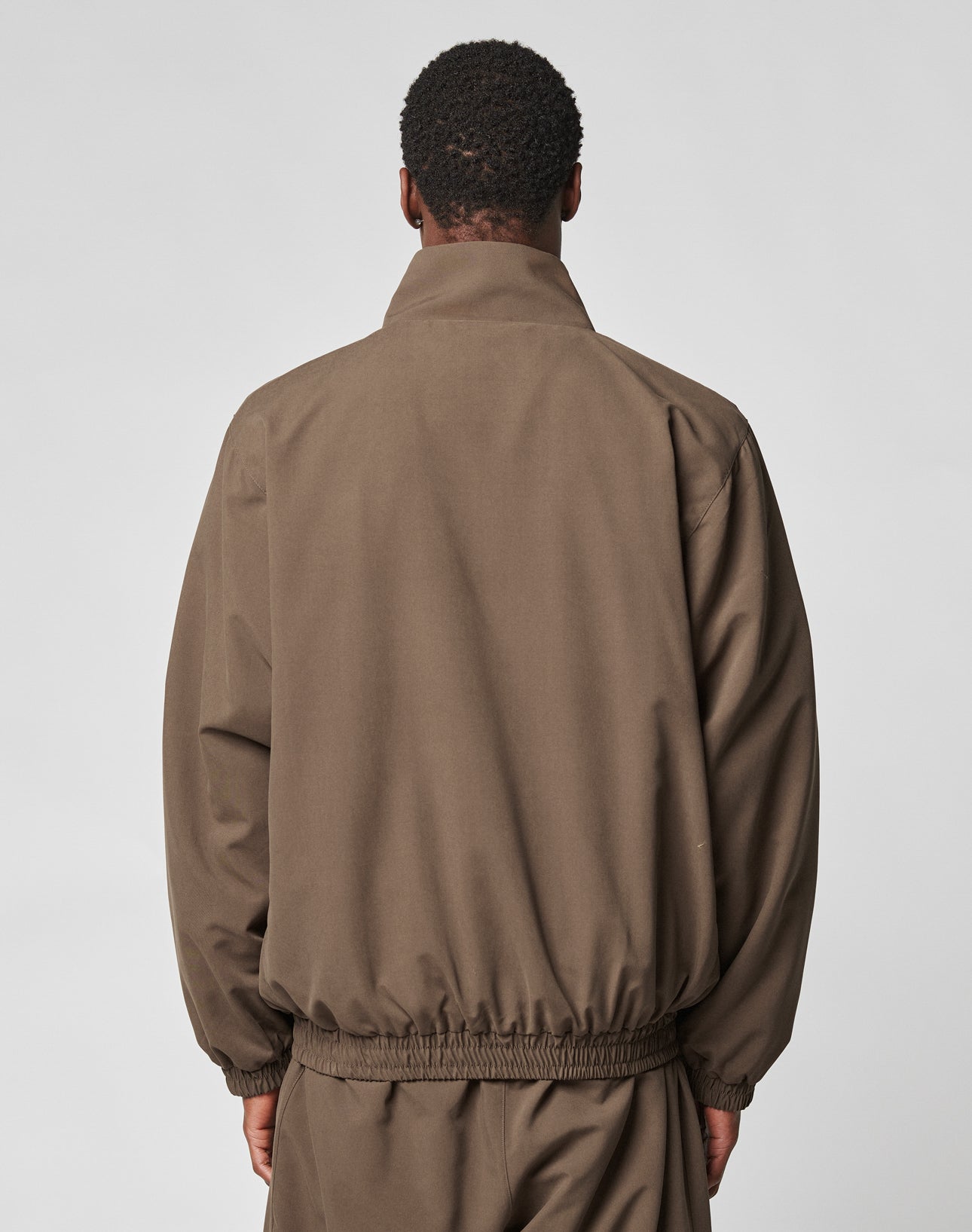 A person with short curly hair, wearing an oversize brown LFDY Trackjacket and matching polyester pants from LIVE FAST DIE YOUNG, faces away from the camera against a plain light gray background.